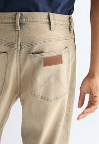 Μπεζ τζιν Wrangler με πίσω τσέπη και δερμάτινο λογότυπο, φορεμένα από άτομο με σκούρο μπλε πουκάμισο και χαλαρό χέρι στο πλάι.