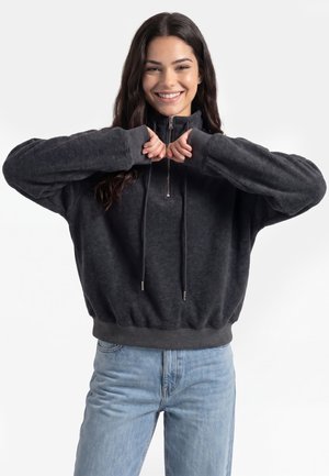 Smilende kvinde i mørk trøje med lynlås og lyseblå jeans står med hævede arme og hænderne foldet sammen ved hagen, vendt fremad.
