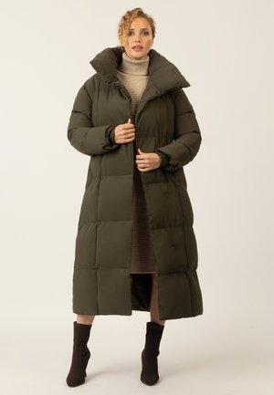 Long dark green puffer coat with oversized collar, quilted design, and snap buttons. Worn over a ribbed brown dress and dark ankle boots.