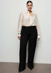 Light beige, sheer button-up shirt with long sleeves paired with high-waisted, straight-leg black trousers. Silver cuff bracelet on the wrist.