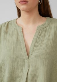 Hellgrüne, strukturierte Bluse mit einem flachen V-Ausschnitt und minimalistischer Kragen. Dezente Falten und gestickte Details in der Nähe des Ausschnitts.