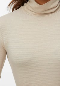 Smėlinis golfas, pagamintas iš raštuoto audinio, su subtilia tekstūra ir prigludusia konstrukcija. Nėra matomų raštų ar detalių.