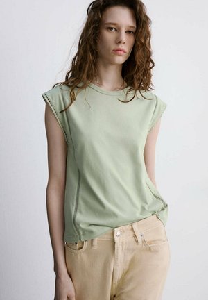 Jeune femme aux cheveux bruns bouclés portant un top sans manches vert clair et un pantalon beige sur un fond blanc uni.