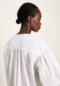 Frau mit geflochtenem Haar in einem Dutt, die eine lockere, weiße Bluse trägt, von hinten und zur Seite gegen einen schlichten, hellen Hintergrund gezeigt.