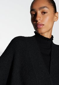 Zwarte gebreide trui met een gestructureerd patroon en een ruches aan de halslijn. Het ontwerp heeft een diepe V-vorm en lange, brede mouwen.