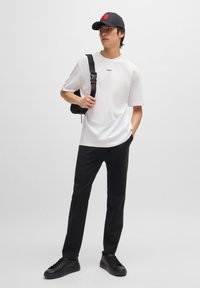 Vit bomullströja med "HUGO"-logotyp, matchad med svarta skräddarsydda byxor och svarta sneakers. Modellen bär en svart axelväska och har på sig en svart keps.