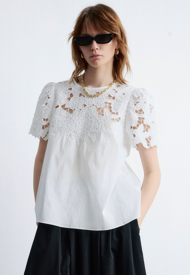 White blouse with lace floral cutouts on shoulders, short sleeves, and a relaxed fit. Paired with a black skirt and gold chain necklace.