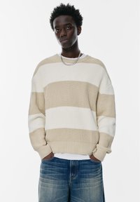 Beige en witte gestreepte gebreide trui met ribbelstructuur, ronde halslijn en verlaagde schouders, gecombineerd met lichtblauwe denim jeans.