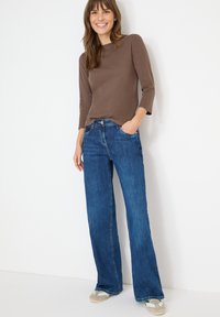 Bruine nauwsluitende top met lange mouwen, gecombineerd met losse blauwe uitlopende jeans. De jeans hebben klassieke zakken en een lichte denimtextuur.