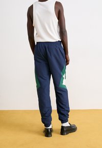 Marina blå byxor för träning med elastiska muddkanter, med gröna och vita geometriska detaljer. Materialet verkar vara lättvikts och har en slät struktur.