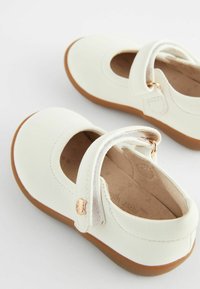 Białe skórzane buty Mary Jane z okrągłym noskiem, brązową gumową podeszwą, regulowanym paskiem i małym złotym zdobieniem z boku.