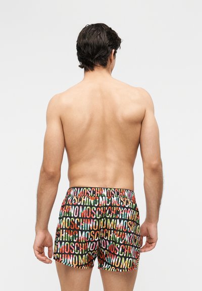 Homme vu de dos portant un short coloré avec un motif répétitif du texte "MOSCHINO" sur un fond uni gris clair.