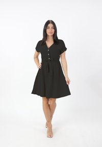 Donna con capelli scuri che indossa un abito nero con bottoni e cintura annodata, in piedi con una mano sui fianchi e tacchi trasparenti su uno sfondo bianco.