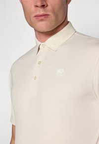 Beige Poloshirt aus weichem Material mit zwei Knöpfen und einem kleinen weißen Logo auf der linken Brust. Kurze Ärmel und klassischer Kragen.