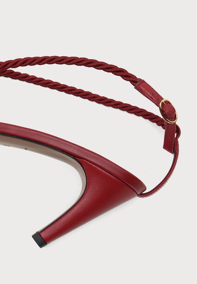 Chaussure à talon haut en cuir rouge avec bride de cheville torsadée et petite boucle dorée sur fond clair.
