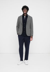 Grå blazer med en texturerad yta, bärs över en mörk skjorta, tillsammans med marinblå byxor och vita sneakers. Har en skräddarsydd passform.