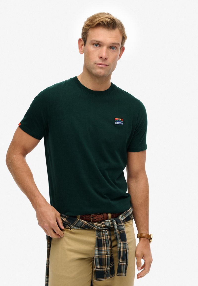 Camiseta de algodón verde con cuello redondo y mangas cortas, que presenta un pequeño parche de logo colorido en el lado izquierdo.