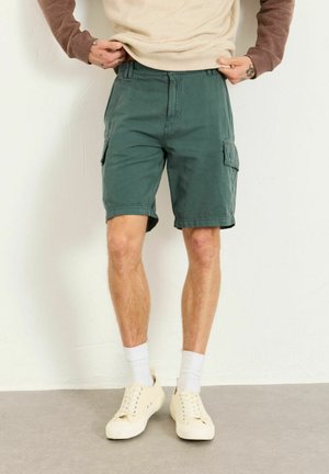 Man wearing green cargo shorts, beige sneakers, white socks, and a beige and brown long-sleeve shirt standing against a white wall.