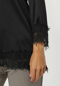 Blusa negra con intrincados detalles de encaje en las mangas y en el dobladillo. Tela suave con una sutil textura. Se combina con pantalones de patrón a cuadros.
