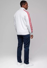 Felpa chiusa leggera grigia con strisce rosse laterali, abbinata a jeans blu scuro e scarpe da ginnastica bianche. Tessuto morbido, vestibilità casual.