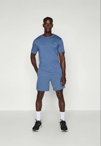 Hombre de pie con las manos en el interior de un atuendo atlético azul, zapatillas negras y calcetines blancos contra un fondo claro y liso.