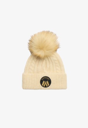 Crèmekleurige gebreide beanie met een gestructureerd patroon, voorzien van een grote nep-bont pompon en een ronde zwarte patch met gouden accenten.