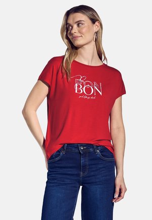 T-shirt rouge à manches courtes avec un col rond, comportant le texte blanc "BON" et "les bonnes choses devraient." Associé à un jean bleu foncé.
