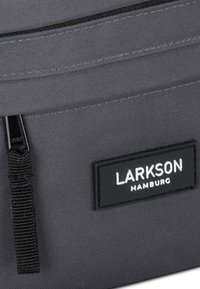 Una borsa in tessuto grigia presenta una superficie liscia, una cerniera nera e un'etichetta rettangolare nera con il testo bianco "LARKSON HAMBURG".