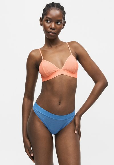 Γυναίκα με πλεξούδες φοράει ένα πλεκτό ροδακινί μπραλέτι και μπλε, ψηλοκαμωμένο ριμπ εσώρουχο, στέκεται με το ένα χέρι στη μέση μπροστά σε άσπρο φόντο.