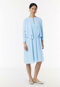 Jasnoniebieska sukienka z plisami i dekoltem w kształcie łezki, w zestawie z dopasowaną otwartą kurtką i beżowymi balerinami. Miękka, zwiewna tkanina.