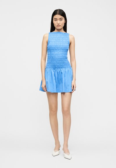 Γυναίκα όρθια, φορώντας ένα μίνι φόρεμα μπλε χωρίς μανίκια με πιέτες και λευκά τακούνια με μυτερή μύτη, μπροστά σε απλό φόντο.