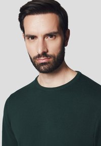 Ciemnozielony sweter z dzianiny z okrągłym dekoltem. Tkanina wygląda na gładką, ma dopasowany kształt i jest noszony przez mężczyznę z przystrzyżoną brodą.