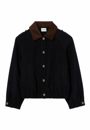 Donna che indossa una giacca nera sopra una felpa con cappuccio a righe marroni e grigie, gonna plissettata blu scuro, calzini crema, scarpe da ginnastica arancioni e tiene una piccola borsa marrone.