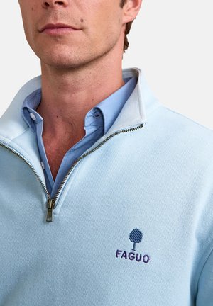 Man wearing light blue quarter-zip sweater over a blue button-up shirt with "FAGUO" logo embroidered on the chest.