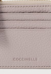 Lähikuva mauve-värisestä nahkastasta, jossa on tikatut korttipaikat ja kohokuvioitu brändi-nimi "COCCINELLE" alaosassa.