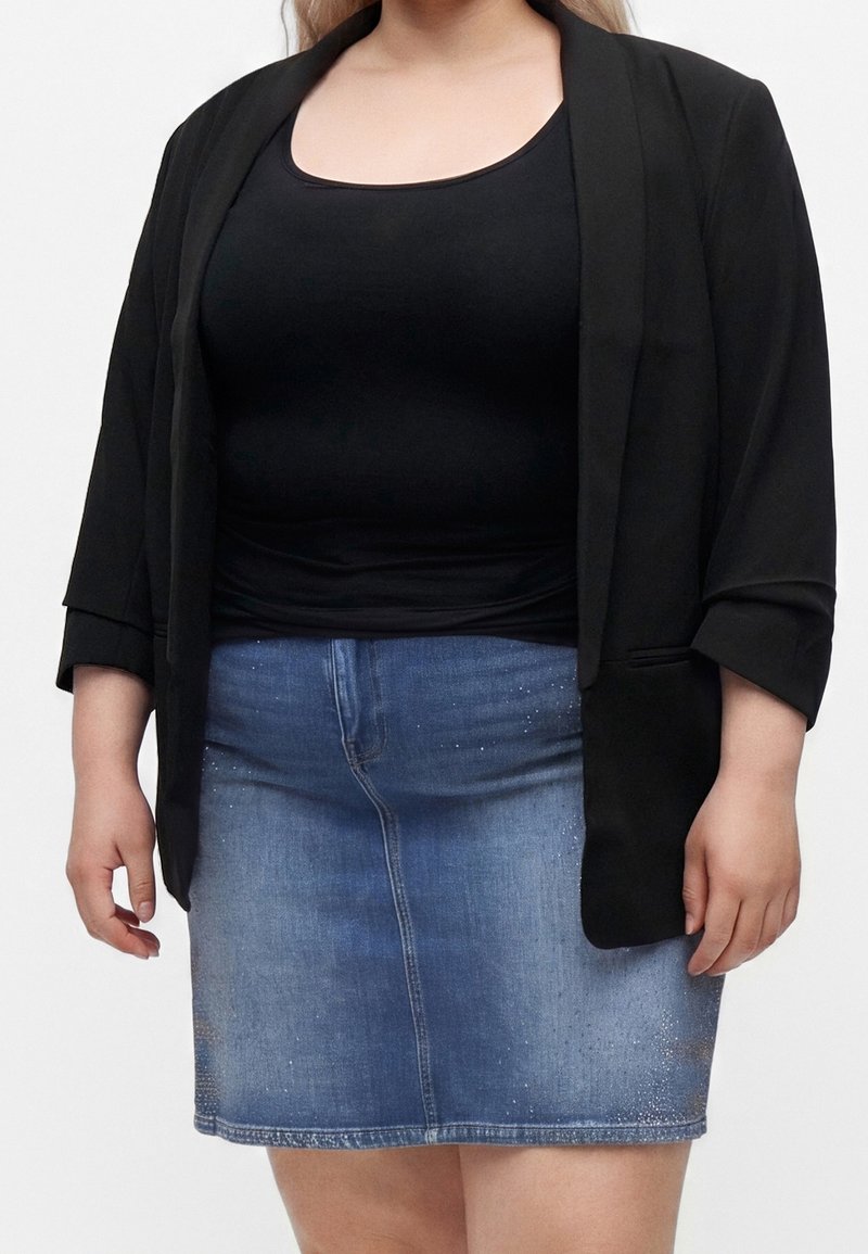 Femme portant un blazer noir et un top associés à une jupe en denim bleu avec des détails subtils scintillants.