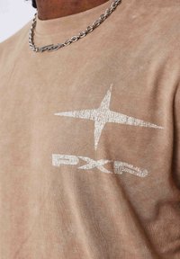 Camiseta de algodón beige con un diseño de estrella blanca texturizada y el logo "PXP" en el área del pecho izquierdo. Se ve una cadena en el cuello.