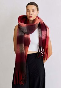 Écharpe en tricot présentant des couleurs dégradées allant du rouge au marron, avec une texture douce et des bords frangés, portée drapée autour du cou.