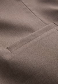 Gros plan d'un tissu marron avec une poche fendue horizontale cousue dans la matière.
