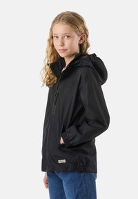 Young girl with long curly blonde hair wearing a black hooded jacket and blue jeans, standing against a plain white background.