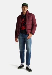 Chaqueta acolchada burdeos con cuello alto, cierre de cremallera y textura acolchada, combinada con una camiseta gráfica negra y jeans azules, zapatos con cordones blancos.