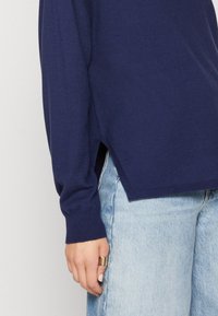 Pull bleu marine à manches longues avec une fente latérale, porté avec un jean bleu clair, avec une bague en or à la main gauche.