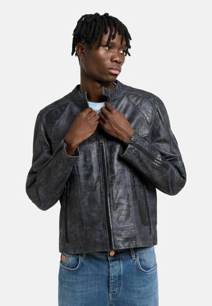 Junger Mann mit kurzen Dreadlocks richtet den Kragen einer schwarzen strukturierten Lederjacke, trägt blaue Jeans und schaut nach rechts vor einem schlichten Hintergrund.
