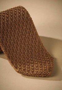 Corbata marrón texturizada con un patrón tejido, que presenta un extremo afilado y una superficie ligeramente elevada. Presentada sobre un fondo neutro.