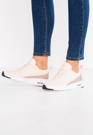 Osoba nosząca jasnoróżowe i beżowe sneakersy Nike z białymi podeszwami oraz niebieskie dżinsy na białym tle.