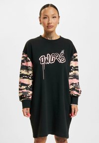 Čierna oversized mikinová šaty s ružovým textom v graffitovom štýle, s rukávmi v ružovom a béžovom maskáčovom vzore a rebrovanými manžetami.