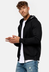 Sort zip-up hoodie med en hvid indvendig foring og logo-accenter på hætten. Har ribbede manchetter og kant, båret over en hvid t-shirt.