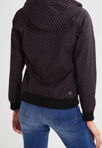 Černá kapucová bunda s červeným puntíkovaným vzorem. Má žebrovaný lem a manžety, s hladkou texturou látky. Nosit s modrými džíny.