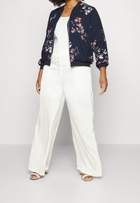 Giubbotto bomber navy con ricami floreali, chiusura zip e polsini elasticizzati, abbinato a pantaloni bianchi a gamba larga e sandali aperti.