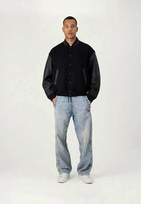 Μαύρο μπουφάν bomber με δερμάτινα μανίκια, συνδυασμένο με ανοιχτό μπλε τζιν παντελόνι και λευκά αθλητικά παπούτσια. Διαθέτει κλείσιμο με κουμπιά και μπροστινές τσέπες.